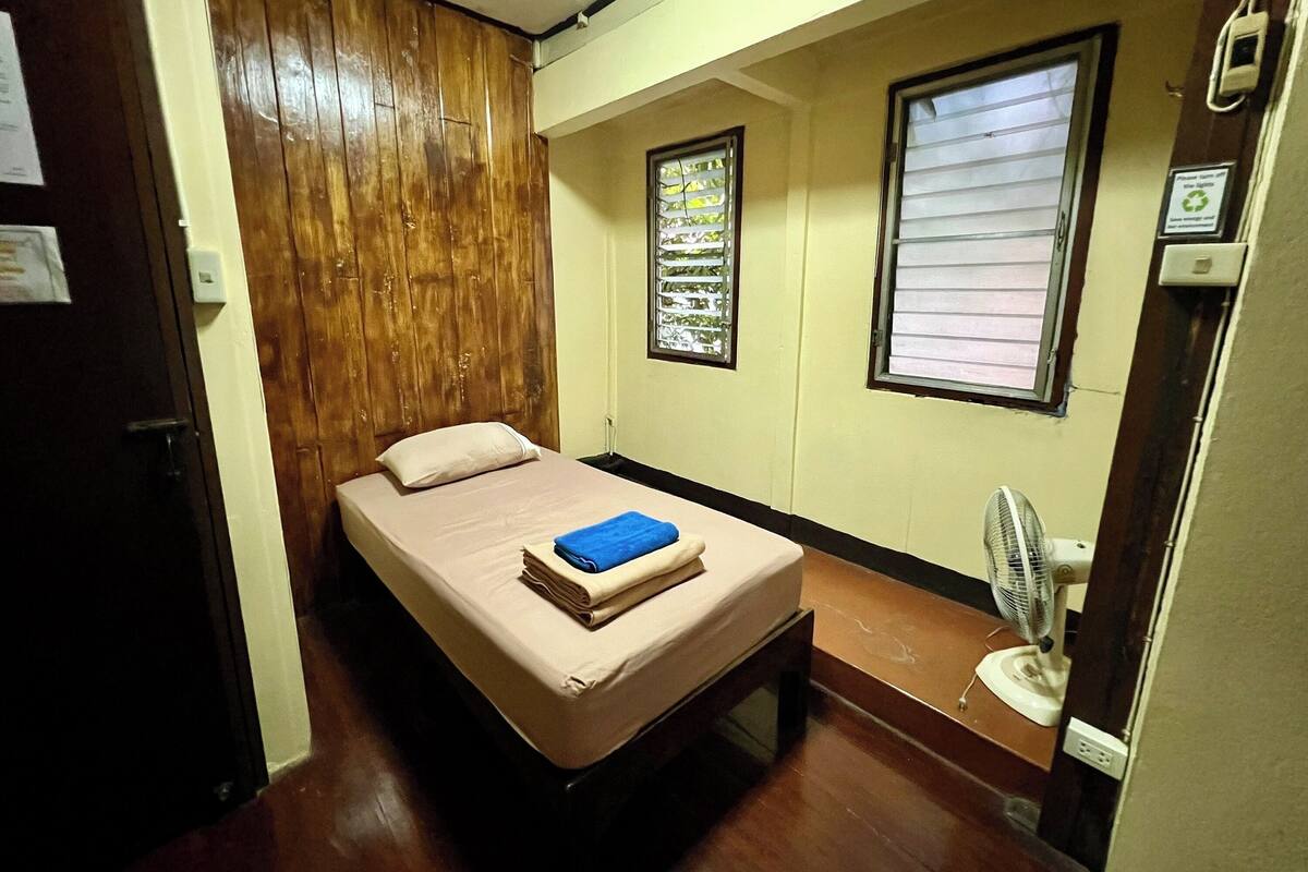 A small, single-bed room with a wooden wardrobe, a window with blinds, and a standing fan.