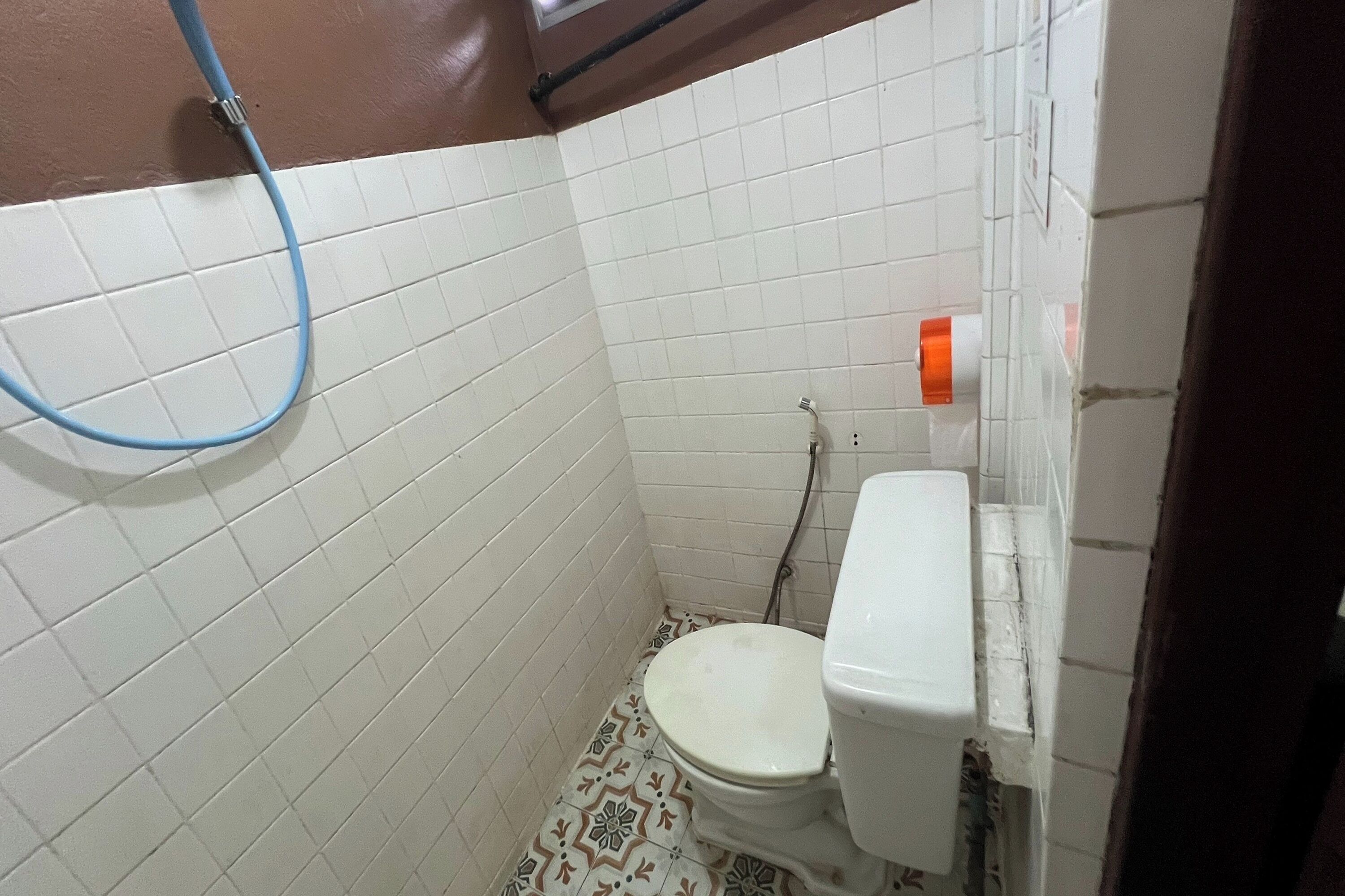 A small bathroom with white tiled walls, a toilet, and a shower hose.
