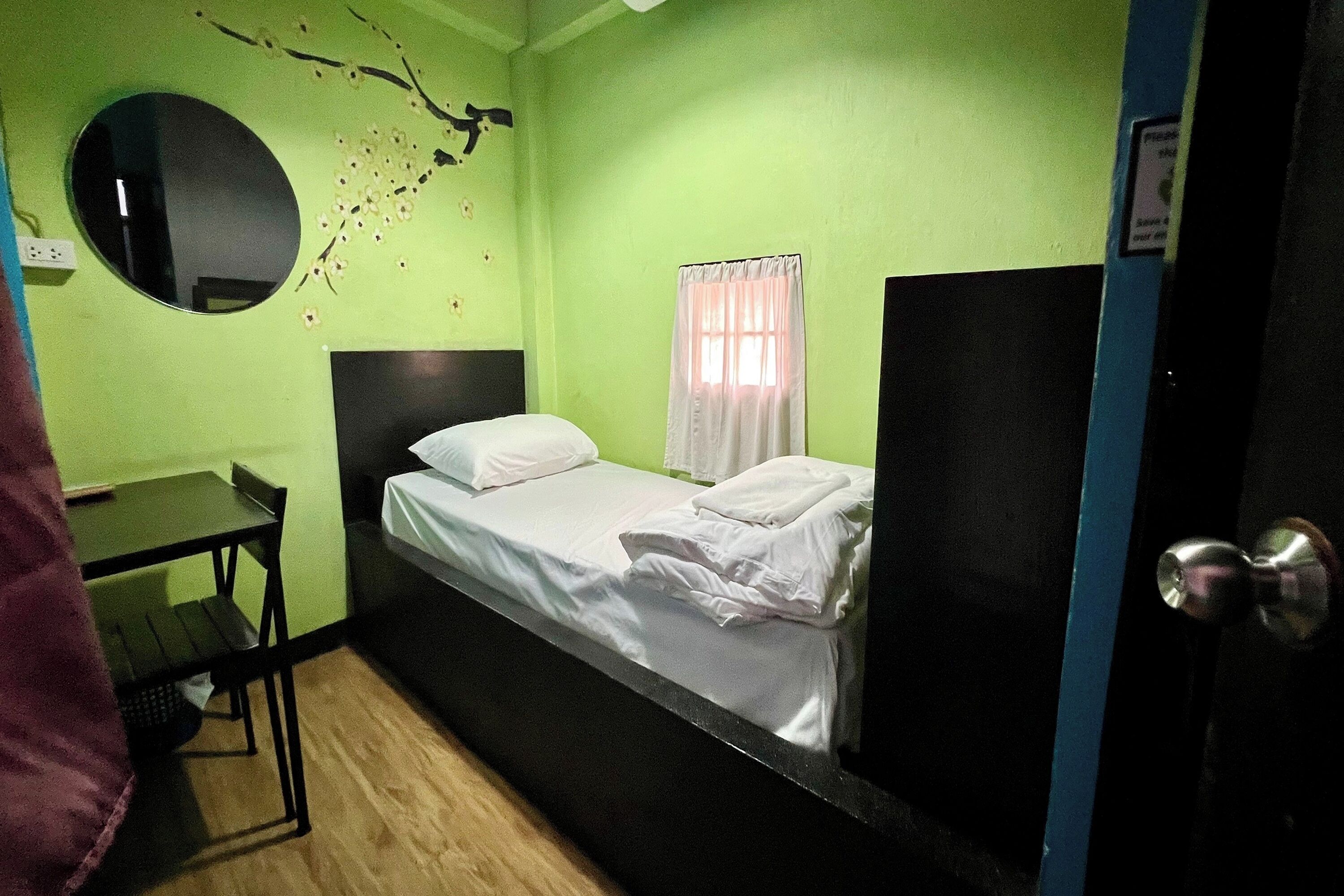 A small, single-bed room with a green wall, a round mirror, a small table, and a window with curtains.