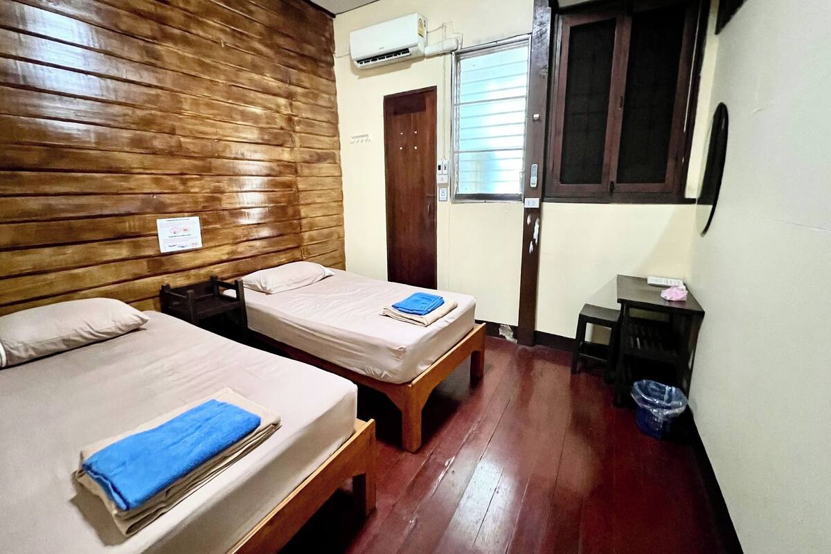A room with two beds, wooden walls, a window with blinds, a door, and a small table with a trash bin.