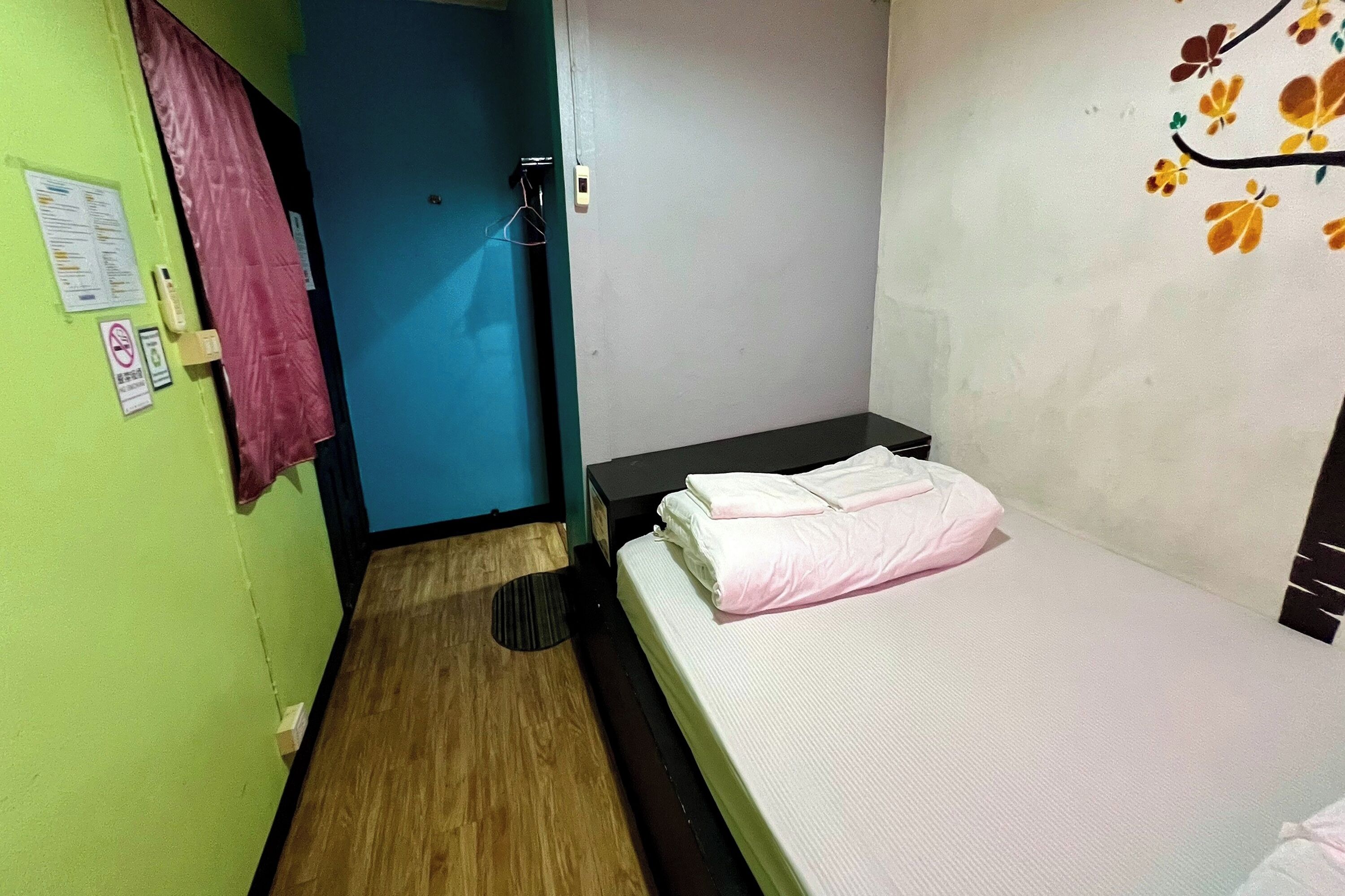 A small, single-bed room with a wooden floor, a blue door, and a wall with a floral mural.