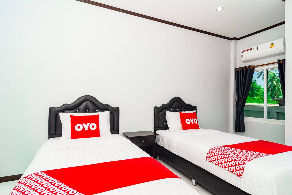 A hotel room with two beds, each with a black headboard and white bedding. There is a window with curtains, an air conditioning unit, and a small table between the beds.