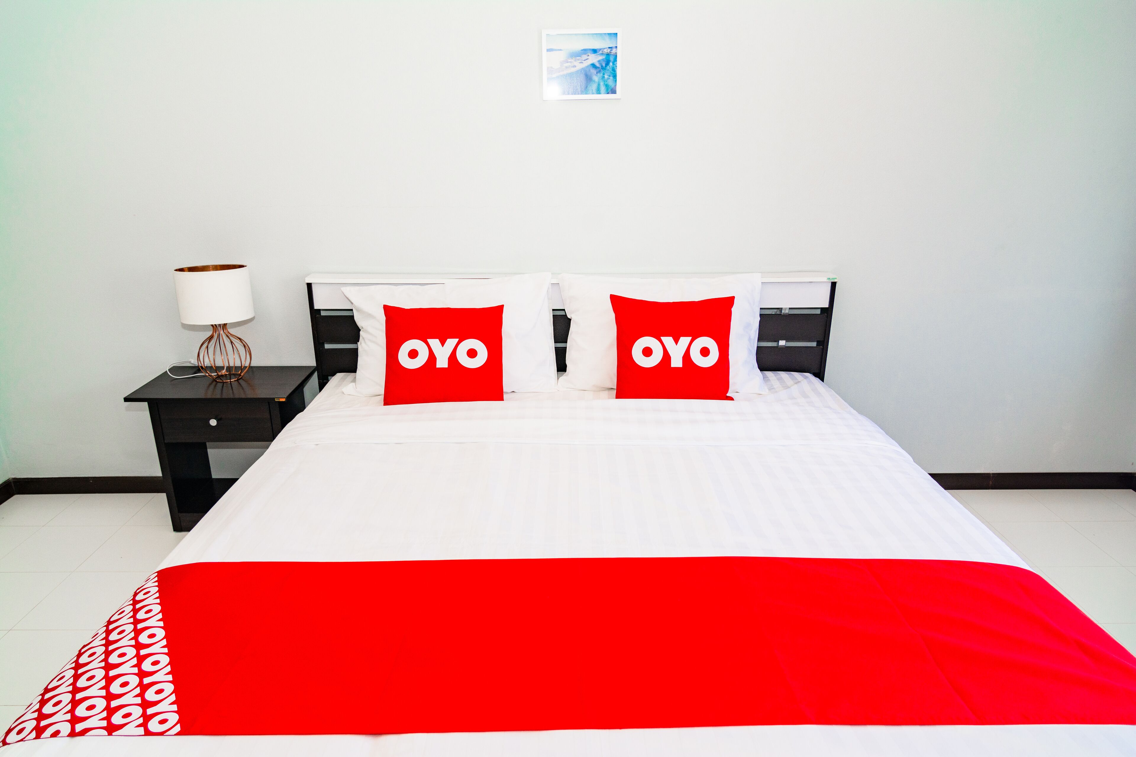 A hotel room with a bed, two pillows with the word "OYO" on them, a nightstand with a lamp, and a picture on the wall.