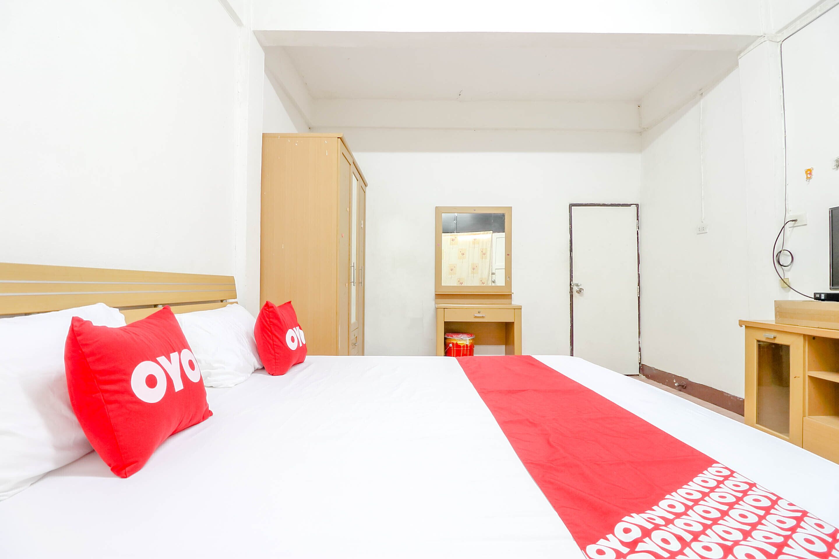 Habitación de hotel con una cama, dos almohadas rojas, un armario de madera, un pequeño mueble de madera, un espejo y un televisor sobre un soporte.