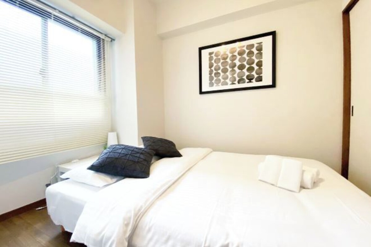 A neatly made bed with white and dark pillows, a framed artwork on the wall, and a window with blinds.
