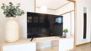 A modern living room with a flat-screen TV on a wooden console, a woven vase with green leaves, and a small potted plant. - OUCHI HOTEL Dambara (Hiroshima)
