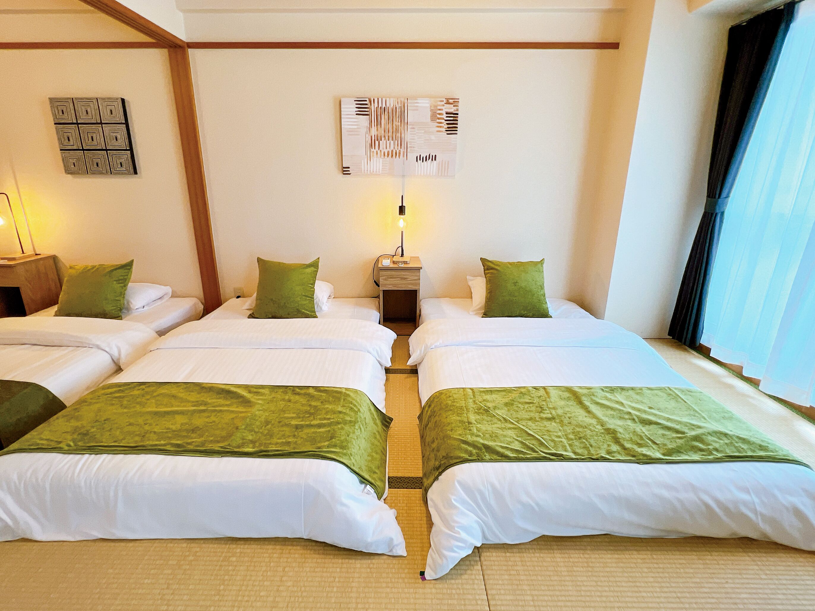 Een hotelkamer met twee bedden, elk voorzien van een groene spreidek en witte lakens, en groene kussens. Daartussen staat een nachtkastje met een lamp, en erboven hangt een kunstwerk aan de muur.