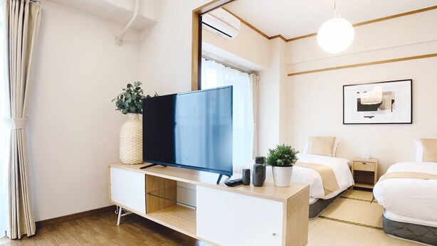 A modern hotel room with a flat-screen TV on a white console, a potted plant, a bedside table with a lamp, a framed picture on the wall, and a large window with curtains. - OUCHI HOTEL Dambara (Hiroshima)