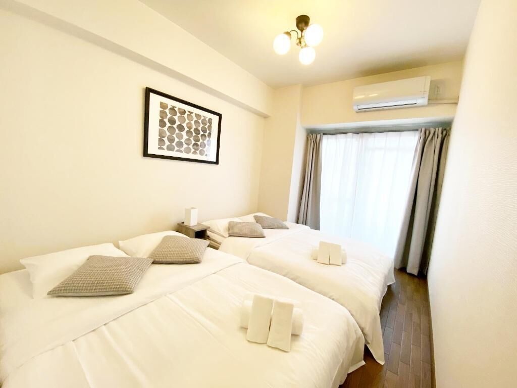 A hotel room with two beds, a framed artwork on the wall, an air conditioning unit, and a window with curtains.