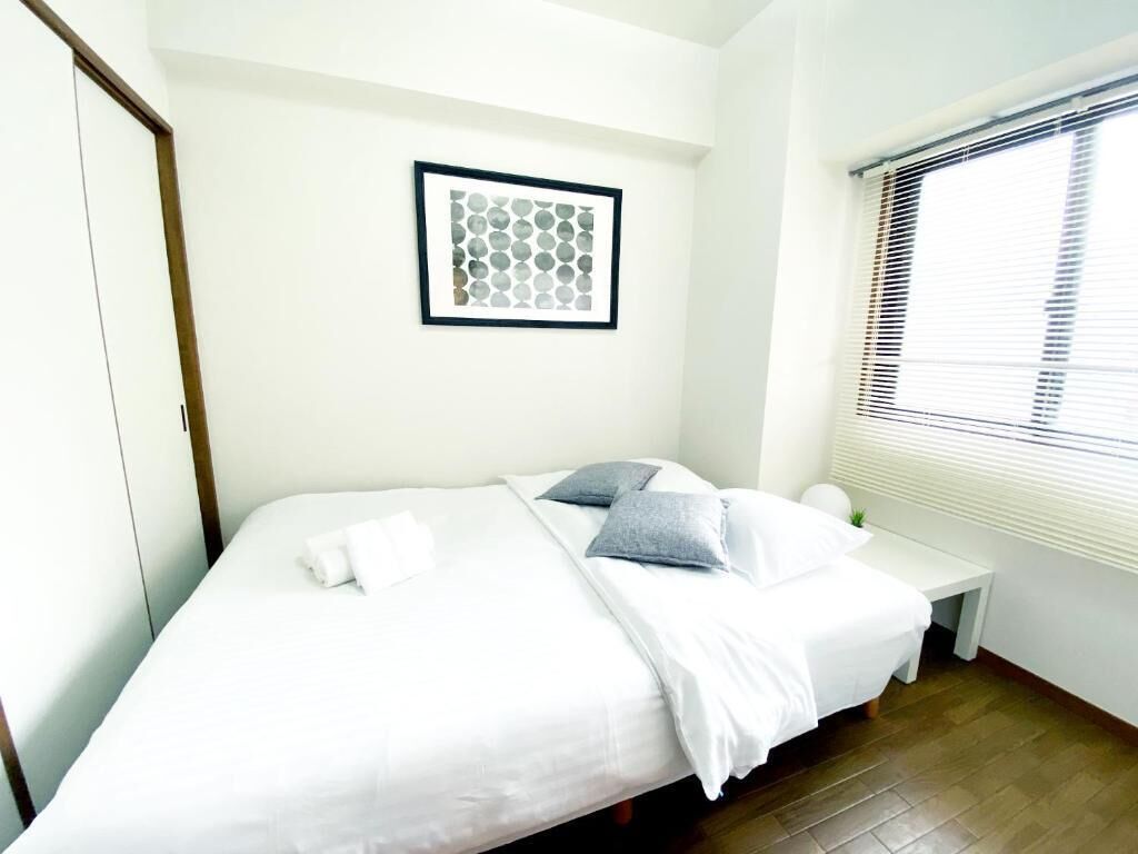 Una cama bien hecha con ropa blanca y dos almohadas, una mesita de noche con lámpara, un cuadro en la pared y una ventana con persianas.