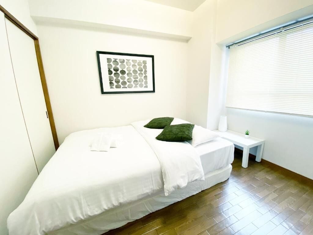 Una cama individual con ropa de cama blanca y almohadas verdes, una mesita de noche con una planta, una obra de arte abstracta enmarcada en la pared y una ventana con persianas.