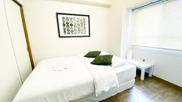 Una cama individual con ropa de cama blanca y almohadas verdes, una mesita de noche con una planta, una obra de arte abstracta enmarcada en la pared y una ventana con persianas.