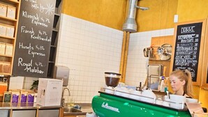 Interior de una cafetería con una máquina de café, una pizarra de menú y una exhibición de cápsulas de café.