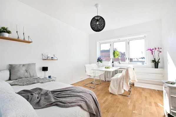 A modern bedroom with a bed, a desk, and a chair. There is a window with a view of buildings outside.