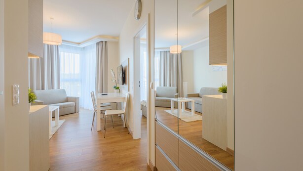 A modern hotel room with a wooden floor, a white sofa, a dining table with chairs, and a large mirror reflecting the room. - Apartamenty NCNK Marina (Sztutowo)