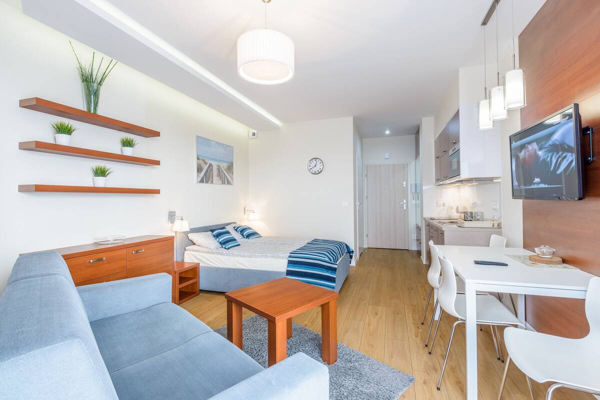 A modern living room with a sofa, coffee table, and a TV. There is a kitchen area with a dining table and chairs.