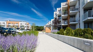 A modern apartment building with a parking lot, a concrete pathway, and purple flowering plants. - Apartamenty NCNK Marina (Sztutowo)