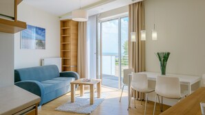 A modern living room with a blue sofa, a wooden coffee table, a white dining table with chairs, and a view of the sea through large windows. - Apartamenty NCNK Marina (Sztutowo)