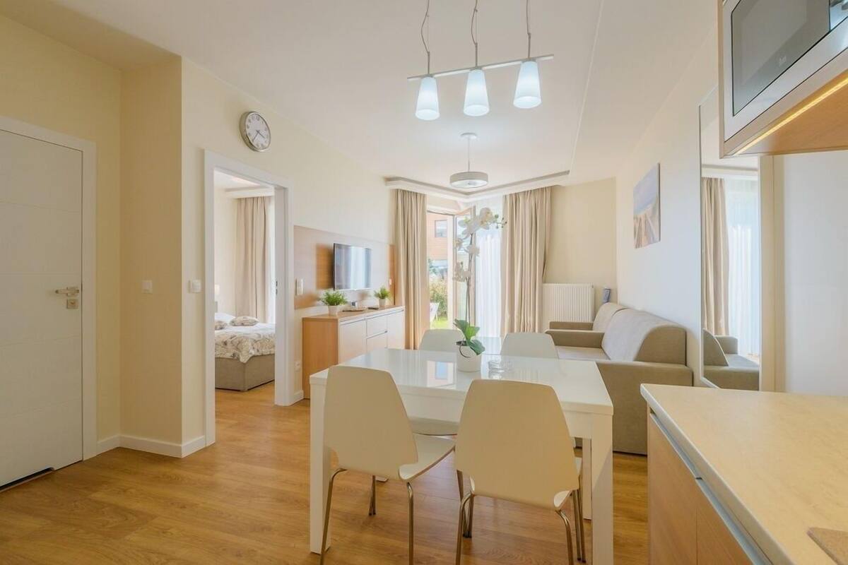 A modern living room with a sofa, a dining table with chairs, a TV mounted on the wall, and a kitchen area with a counter and cabinets.