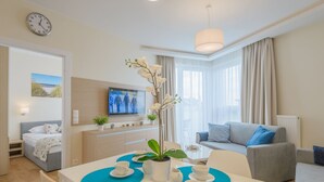A modern living room with a sofa, a dining table with blue placemats, a TV, and a large window with curtains. - Apartamenty NCNK Marina (Sztutowo)