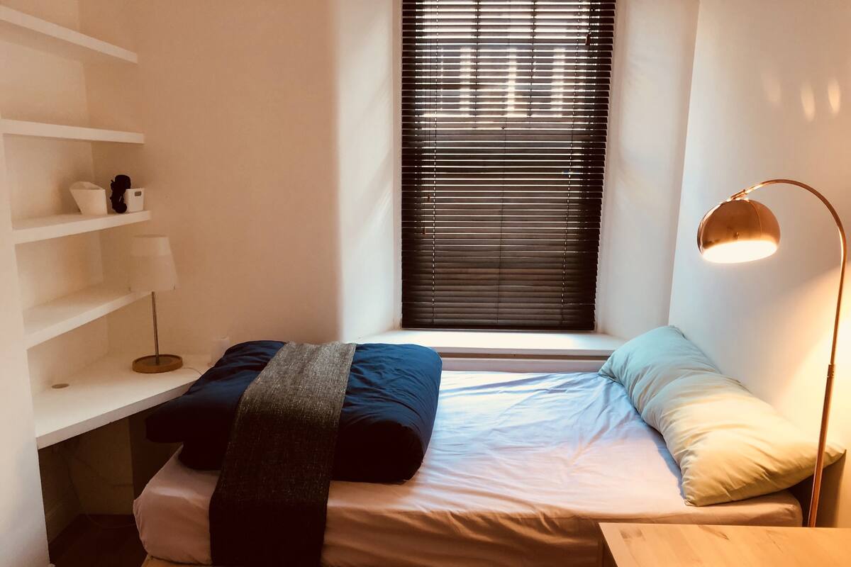 A small, well-lit room with a bed, a window with blinds, a bedside table, and a shelf with a lamp and a few items.