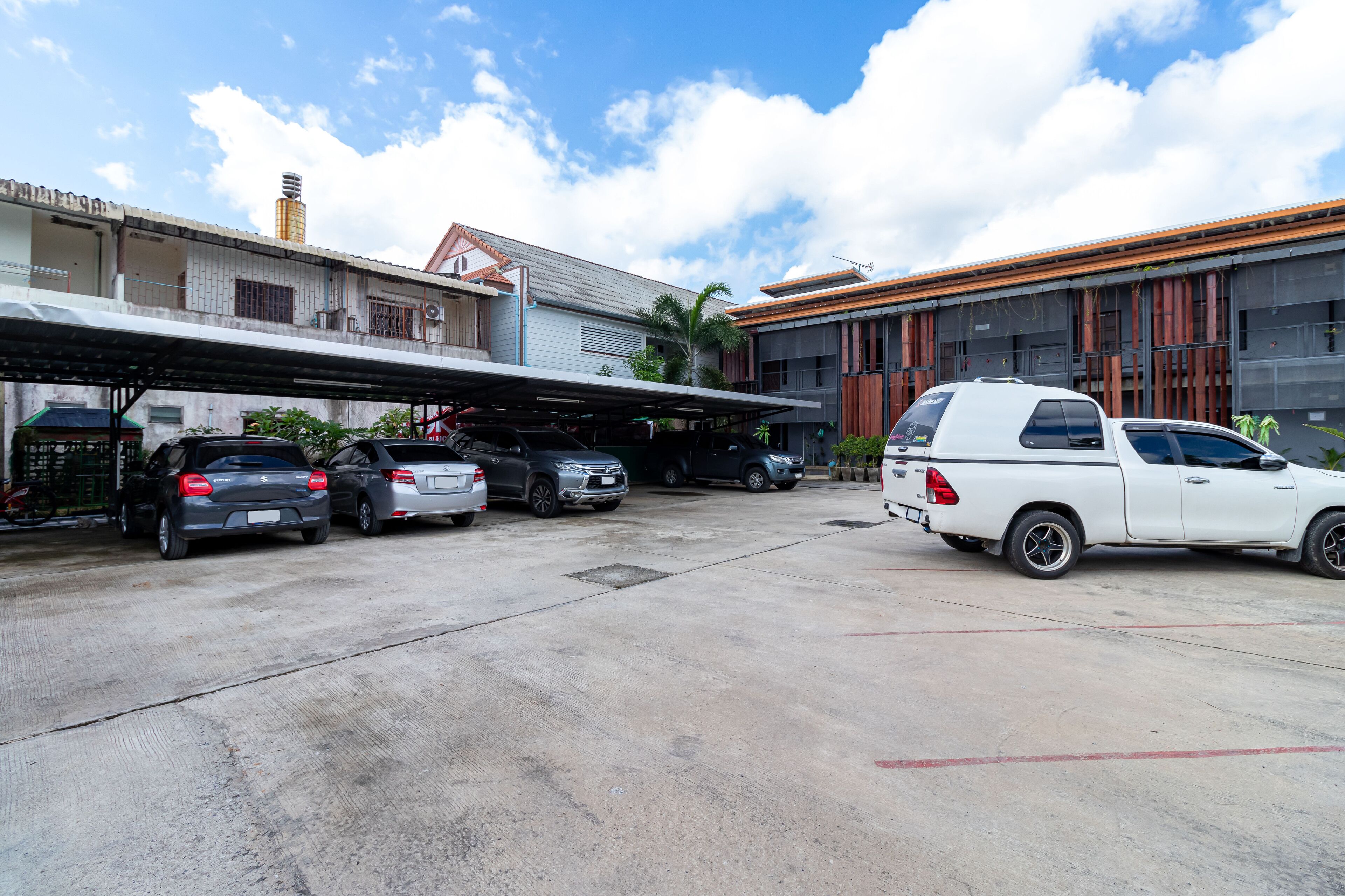 Un aparcamiento con varios coches, incluyendo una camioneta pickup blanca, y un edificio con fachada de azulejos y una sección cubierta.