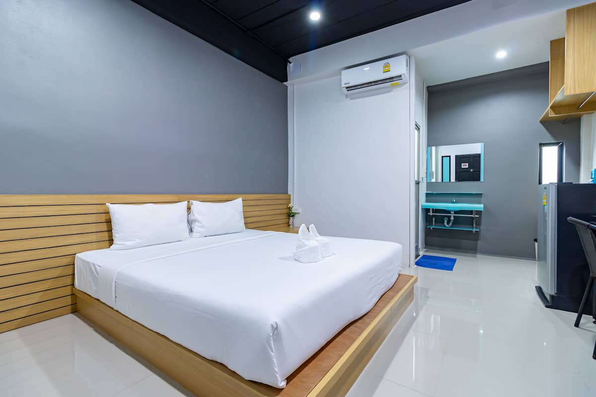 A modern hotel room with a wooden headboard, a large bed with white linens, a built-in air conditioning unit, a small bathroom with a mirror and sink, and a blue mat on the floor.