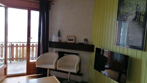 A room with a wooden table, two white chairs, a TV mounted on the wall, a small shelf with a picture frame and a plant, and a sliding glass door leading to an outdoor area. - IRIS Apartment "Vacation Rental in Veysonnaz, 4 Valleys" (Veysonnaz)