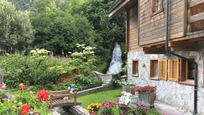 A rustic wooden house with a stone wall, a small garden with flowers, a wooden bridge, and a waterfall. - IRIS Apartment "Vacation Rental in Veysonnaz, 4 Valleys" (Veysonnaz)