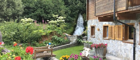 A rustic wooden house with a stone wall, a small garden with flowers, a wooden bridge, and a waterfall.