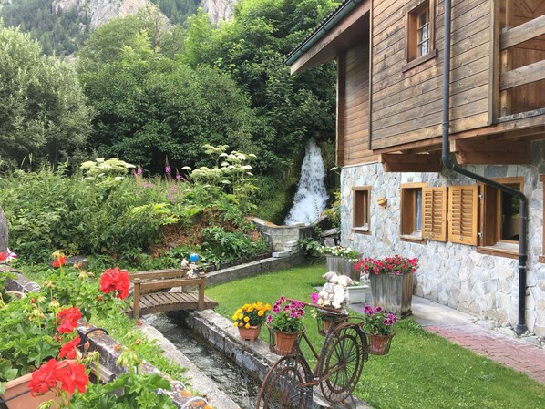 A rustic wooden house with a stone wall, a small garden with flowers, a wooden bridge, and a waterfall. - IRIS Apartment "Vacation Rental in Veysonnaz, 4 Valleys" (Veysonnaz)