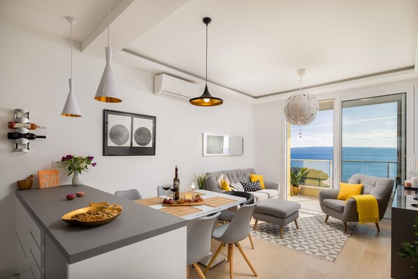 A modern kitchen with a dining area, a grey sofa, and a view of the sea. - Sun Spalato Sky (Split)