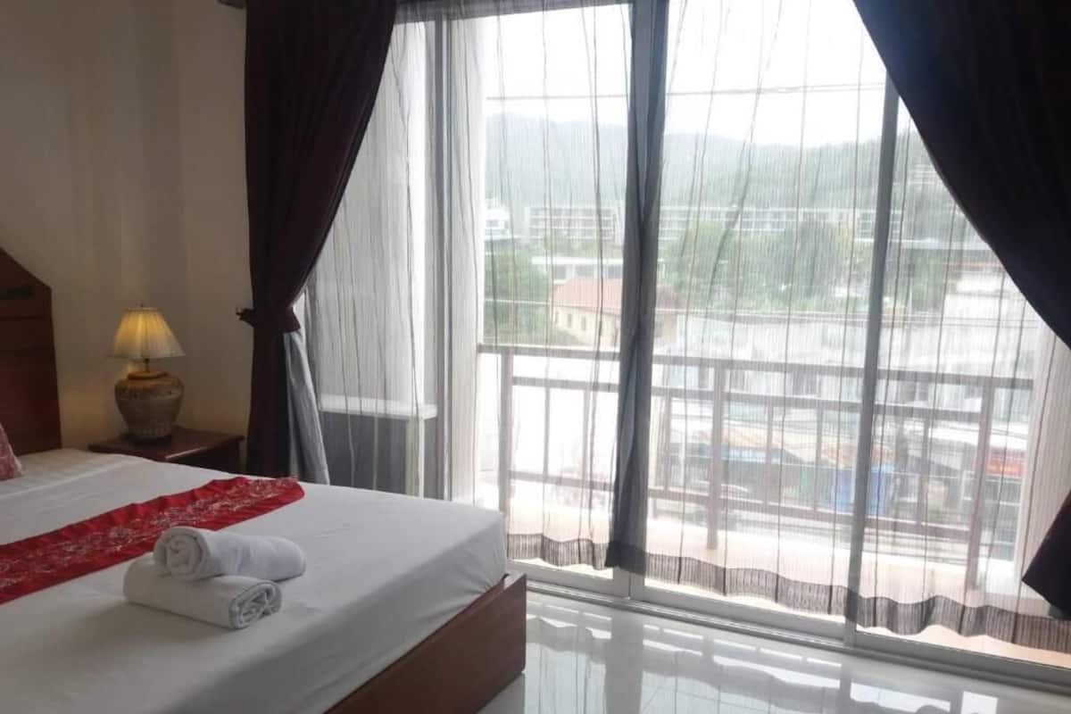 A hotel room with a bed, a lamp, a window with curtains, and a view of buildings outside.