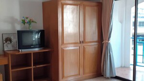 A room with a wooden cabinet, a television on a wooden stand, and a window with curtains. - Praew Mansion (Kamala)