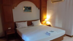 A hotel room with a large bed, wooden headboard, a nightstand with a lamp, and a painting on the wall. - Praew Mansion (Kamala)