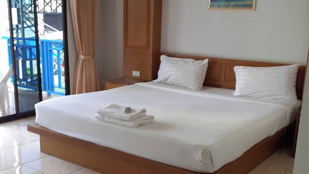 A large bed with white linens and pillows, a wooden headboard, and a small bedside table with a folded white towel. - Praew Mansion (Kamala)