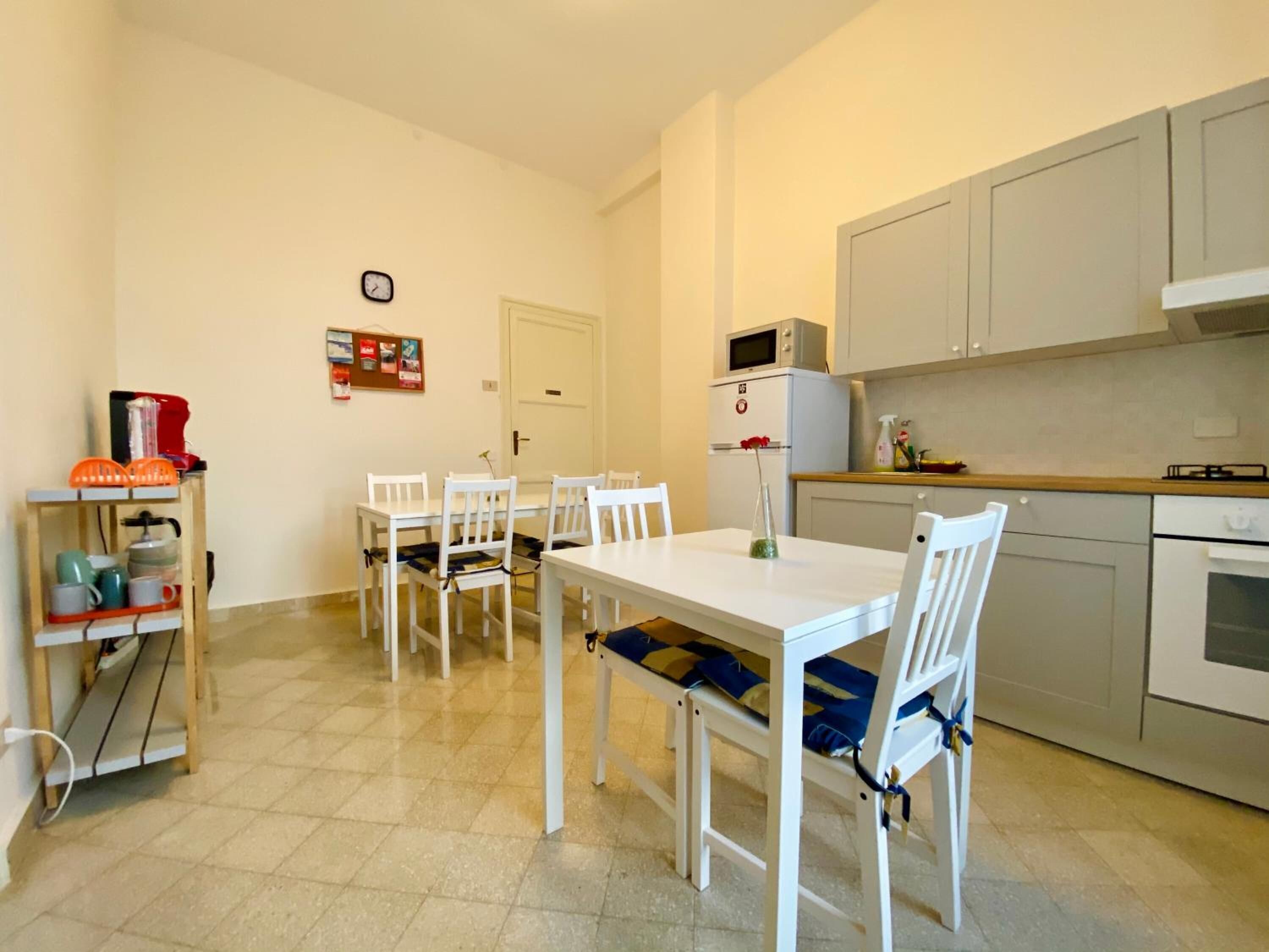 A compact kitchen with white cabinets, a microwave, and a stove. There is a dining table with chairs, a small shelf with kitchen items, and a door with a bulletin board.