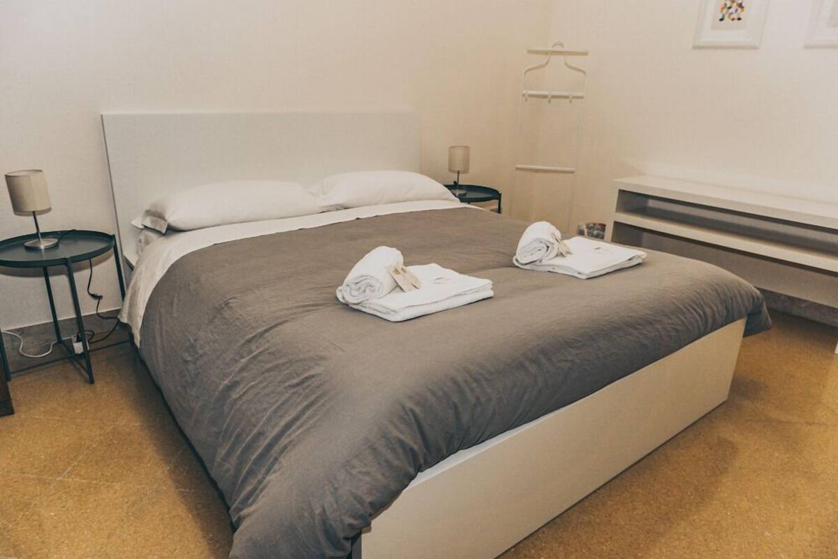 Una cama bien hecha con una colcha gris, dos toallas enrolladas y una mesita de noche con una lámpara.