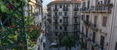 A narrow urban street with multi-story buildings, balconies with plants, and a central open square.