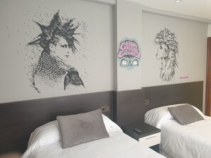 A hotel room with two beds, each with a grey pillow and a remote control on the nightstand.
