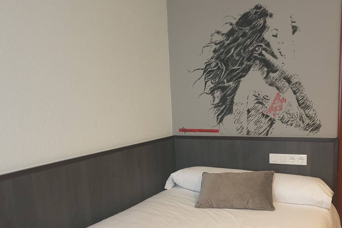 Habitació d'hotel amb un llit, roba de llit blanca, un panell de capçalera gris i una obra d'art a la paret.