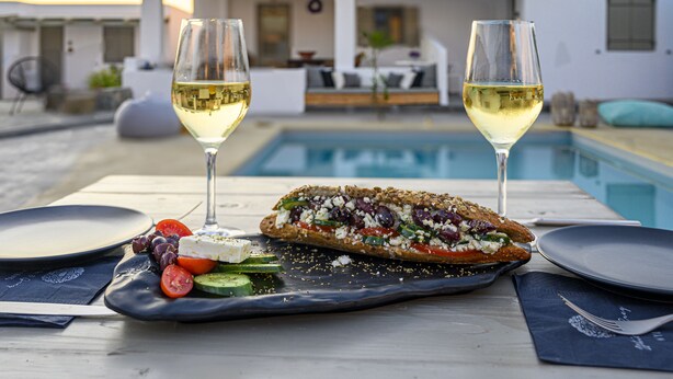 A dining table with a plate of food, two glasses of white wine, and a pool in the background. - White and Grey (Mykonos)