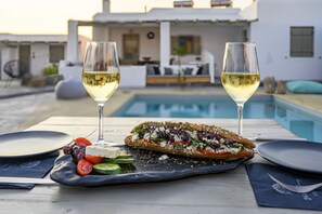 A dining table with a plate of food, two glasses of white wine, and a pool in the background. - White and Grey (Mykonos)