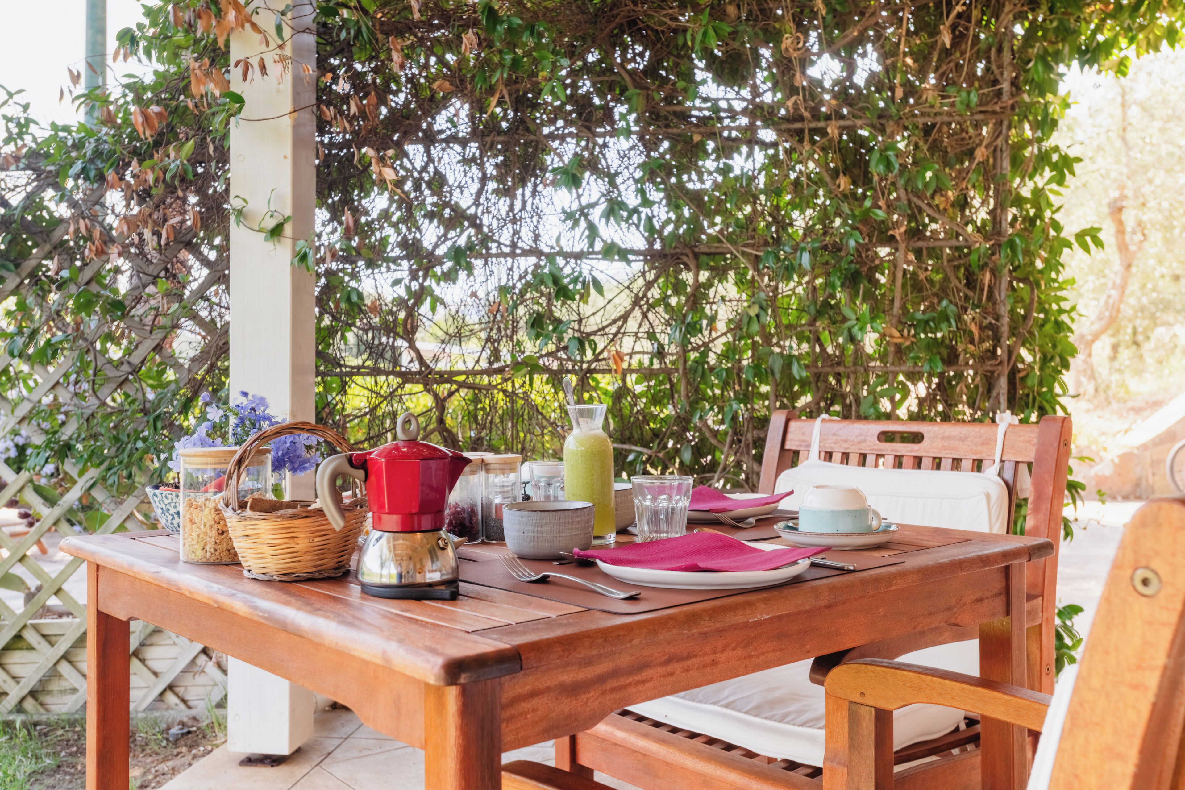 Un área de comedor al aire libre con una mesa de madera, sillas y varios objetos como un hervidor, cuencos y una cesta.