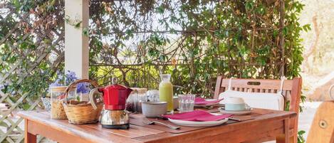 Un área de comedor al aire libre con una mesa de madera, sillas y varios objetos como un hervidor, cuencos y una cesta.