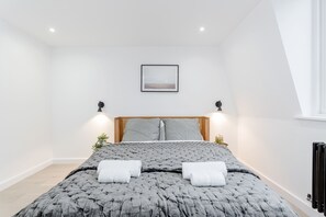 A modern bedroom with a large bed, a wooden headboard, two wall-mounted lamps, a framed picture above the bed, and a radiator on the right.