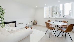 A modern living room with a white sofa, a dining table with chairs, a radiator, and a large window with a view of buildings. - Perfect Holiday Escape - 1 and 2 Bedroom Deluxe Apartments at Liverpool Street (London)