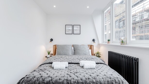 A modern bedroom with a large bed, two bedside lamps, a radiator, and a window with a city view. - Perfect Holiday Escape - 1 and 2 Bedroom Deluxe Apartments at Liverpool Street (London)