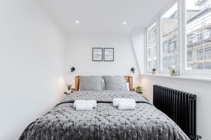 Une chambre moderne avec un grand lit, deux lampes de chevet, un radiateur et une fenĂŞtre donnant sur la ville.