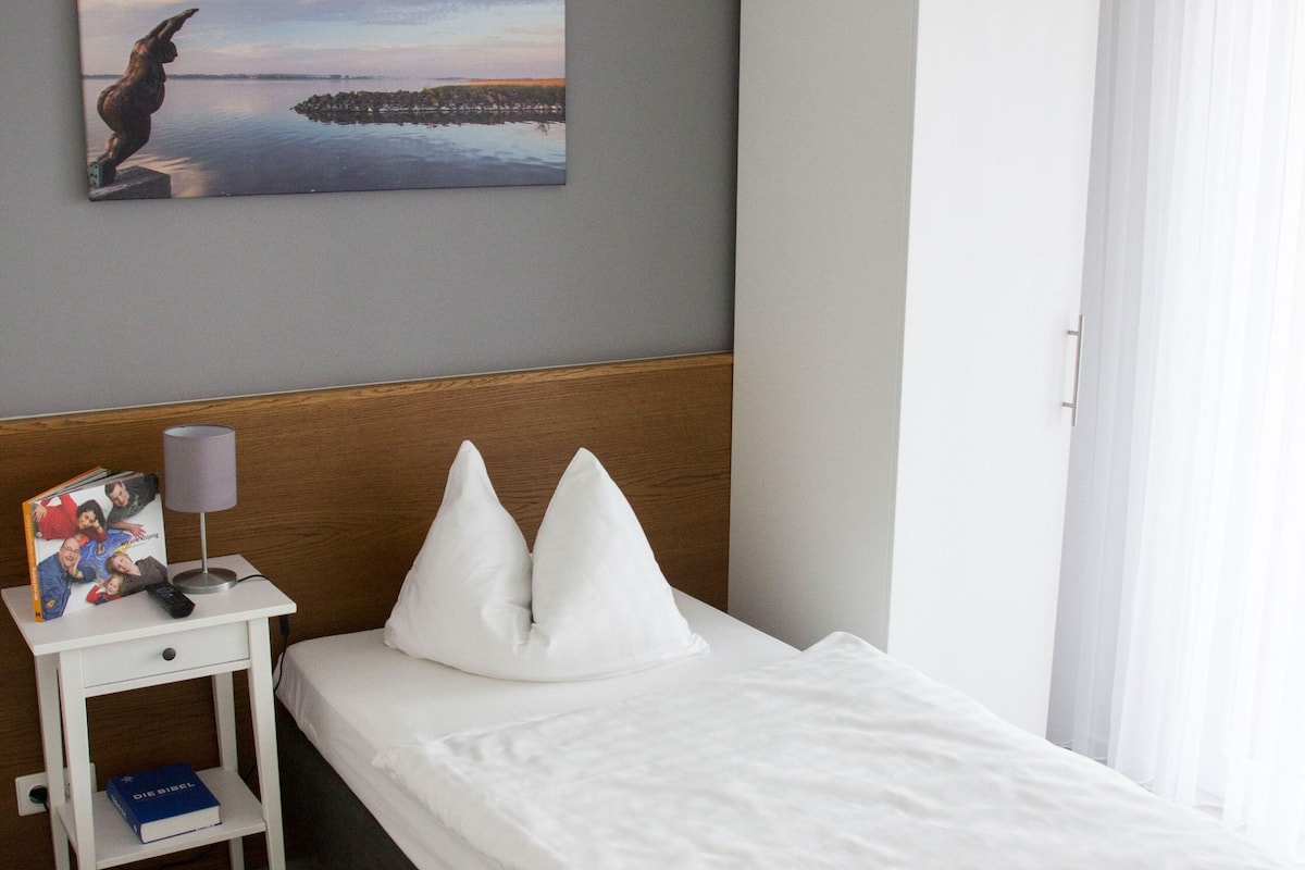 Een hotelkamer met een bed, nachtkastje, een witte kledingkast, een schilderij aan de muur en een raam met gordijnen.